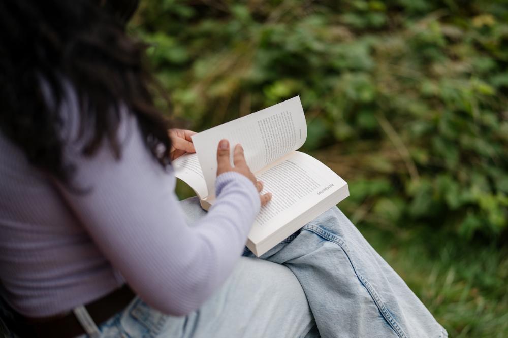 The camera looks over the shoulder of a woman sitting on a bench and reading a book.